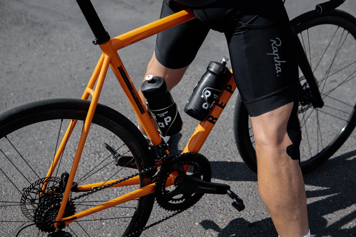 Close-up view of cyclist pedaling matte orange carbon road bike with dual bottle cages mounted on pavement